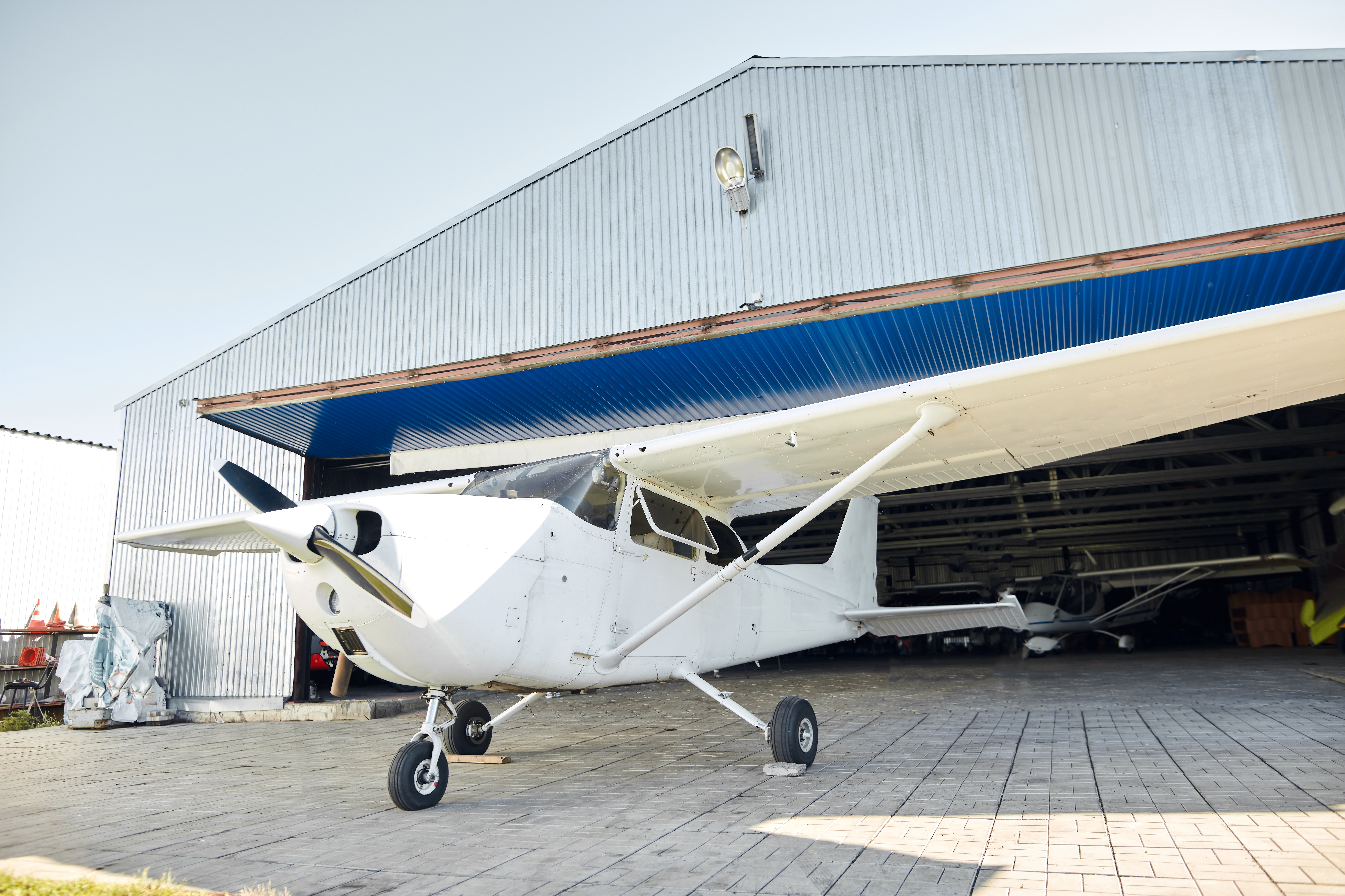 Small white single-engine airplane parked on paved ground before an open corrugated metal hangar.